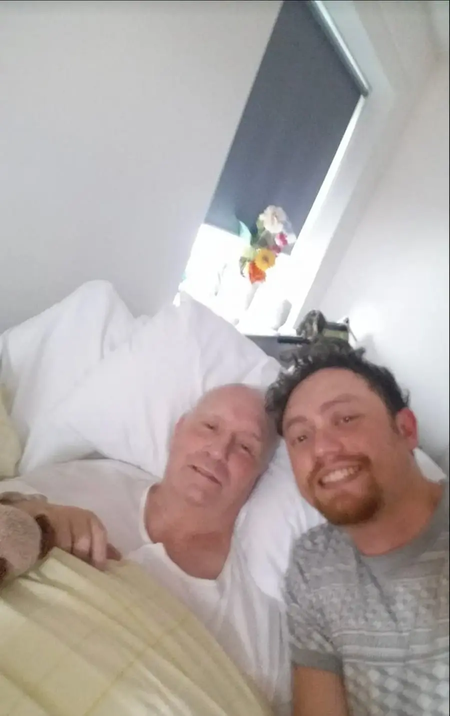 The artist smiling beside his father, who lies in bed with flowers on the windowsill behind them