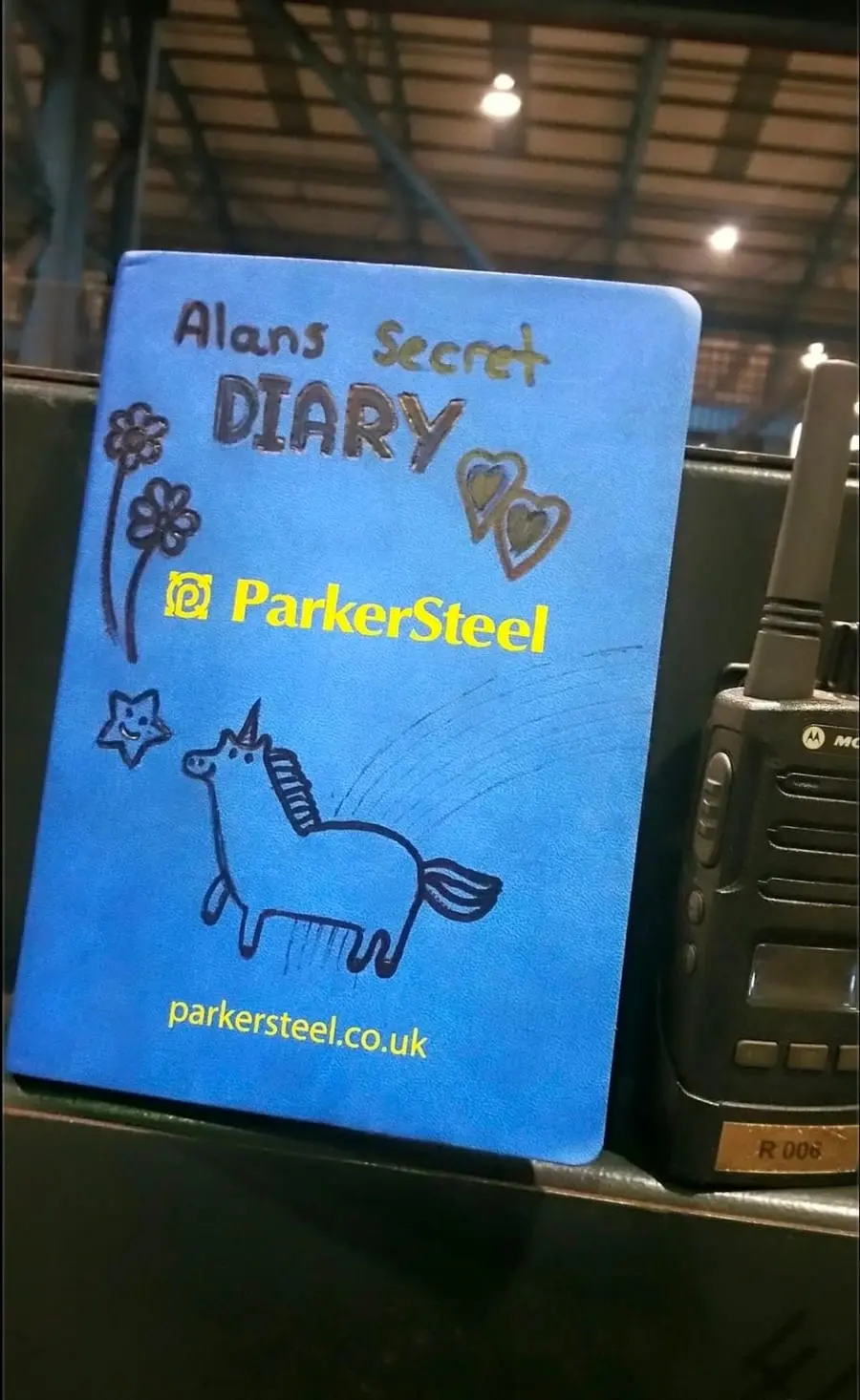A blue ParkerSteel notebook doodled with flowers, hearts and a unicorn, titled Alans Secret Diary, beside a factory radio