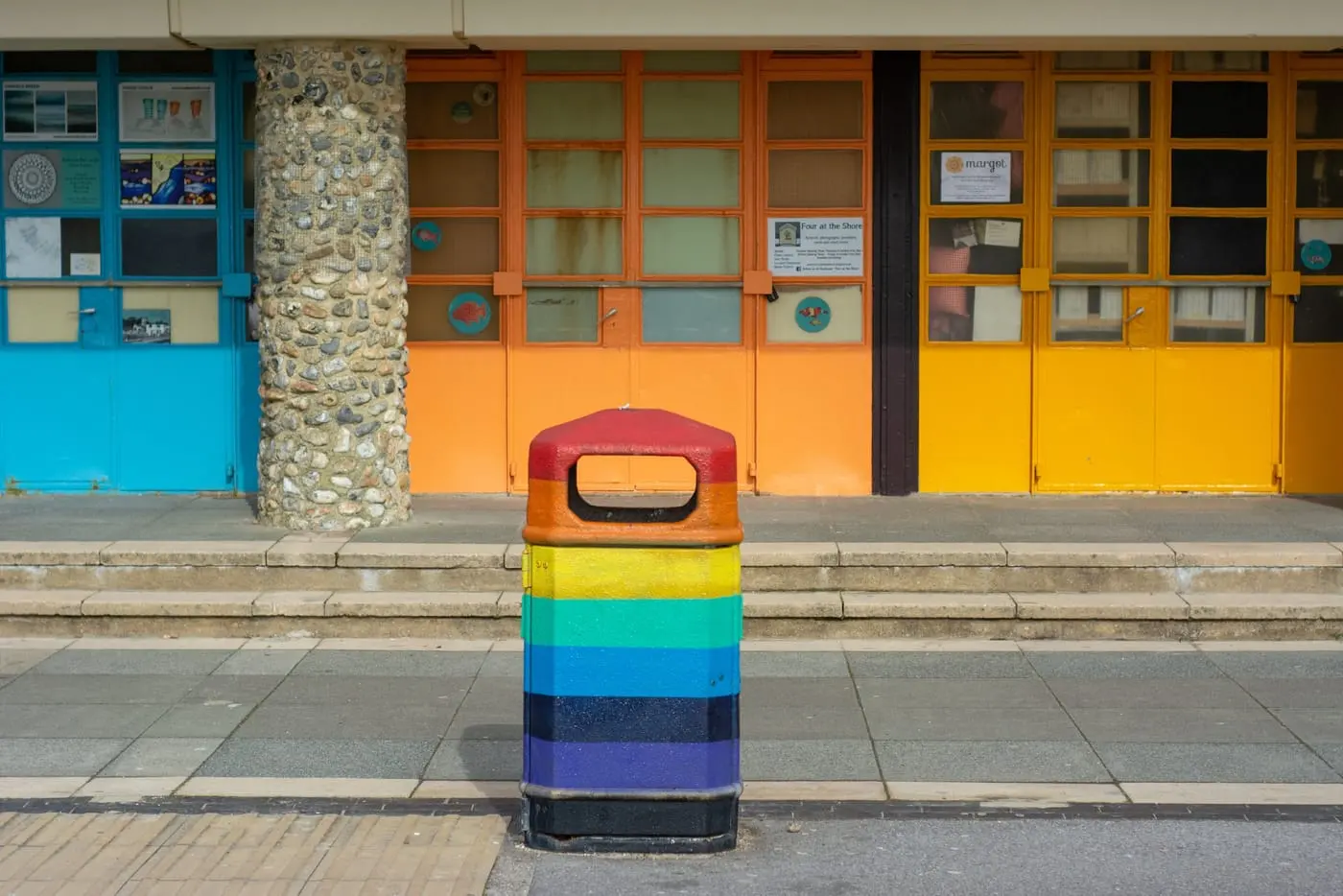 Photography — a rainbow-painted litter bin in front of bright orange and blue seafront shutters